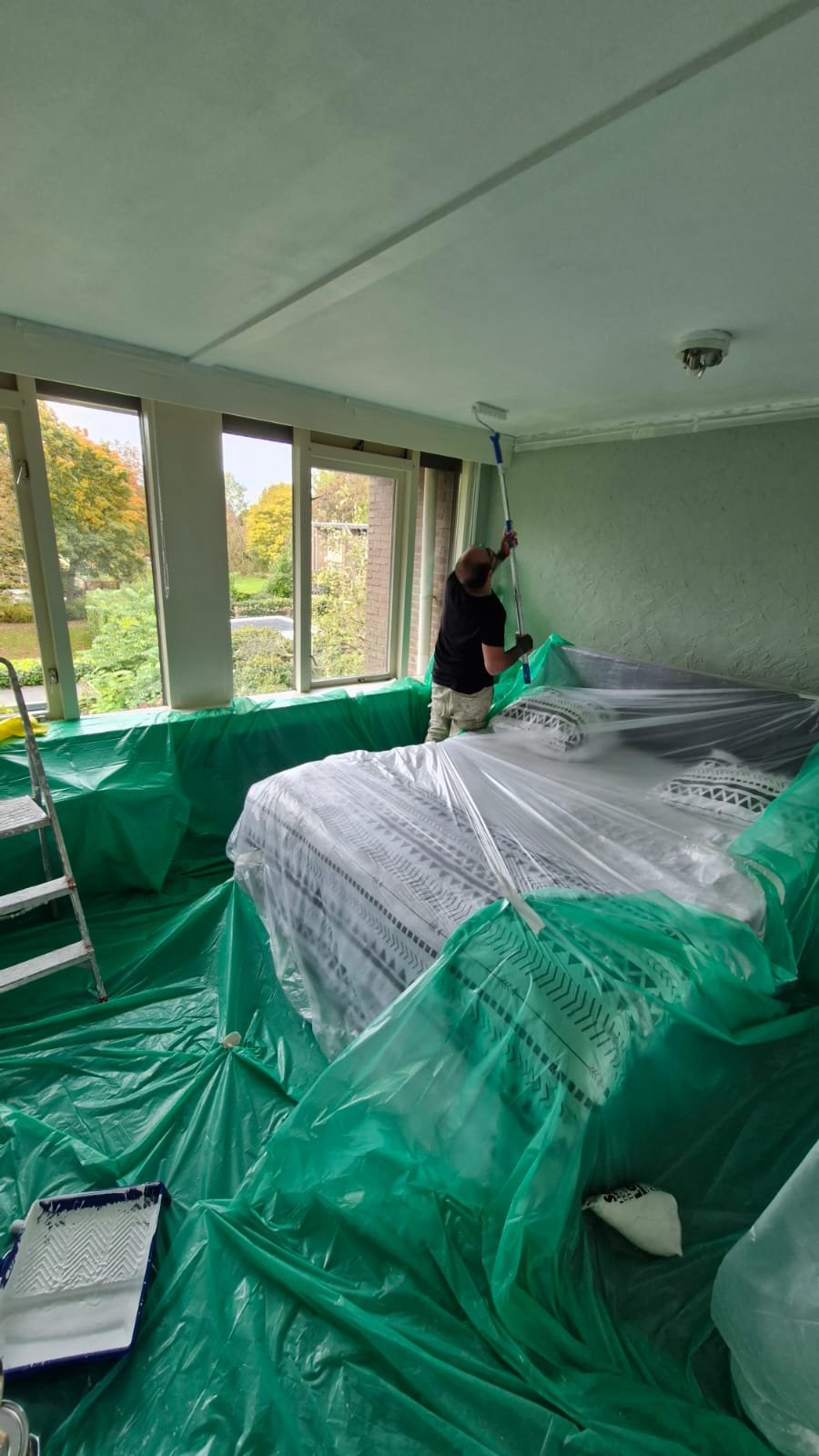 Schilder verft het slaapkamerplafond met een verfroller boven afgedekte meubels